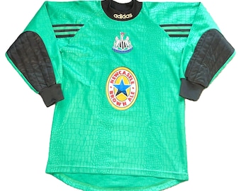 May include: A bright green long-sleeve goalkeeper jersey featuring a black crocodile-skin pattern, black padded elbows, and black cuffs. The shirt displays the Adidas logo, the Newcastle United crest, and a Newcastle Brown Ale graphic.