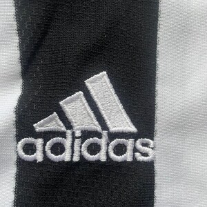 Puede incluir: Primer plano de una tela a rayas blancas y negras con el logotipo de Adidas bordado en blanco. El logotipo presenta tres rayas inclinadas y el nombre de la marca en una fuente sans-serif.