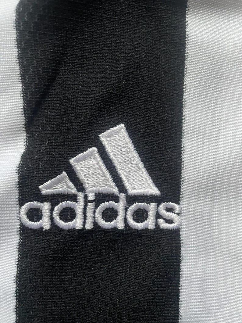 Puede incluir: Primer plano de una tela a rayas blancas y negras con el logotipo de Adidas bordado en blanco. El logotipo presenta tres rayas inclinadas y el nombre de la marca en una fuente sans-serif.