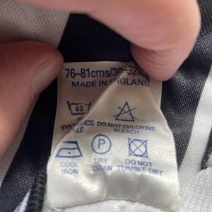 Op de afbeelding: Een wit label met zwarte tekst en verzorgingssymbolen. Het label luidt "76-81cms/30-32in" en "MADE IN ENGLAND". Verzorgingssymbolen omvatten instructies voor wassen, strijken en chemisch reinigen.