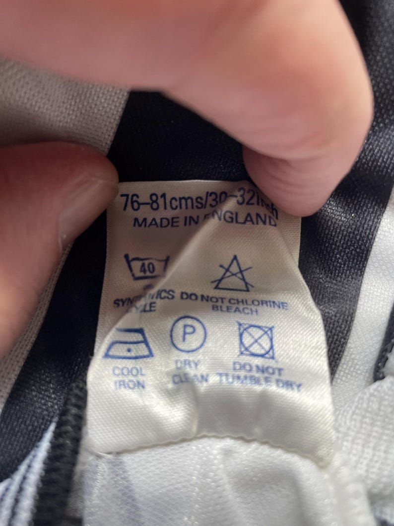 Op de afbeelding: Een wit label met zwarte tekst en verzorgingssymbolen. Het label luidt "76-81cms/30-32in" en "MADE IN ENGLAND". Verzorgingssymbolen omvatten instructies voor wassen, strijken en chemisch reinigen.