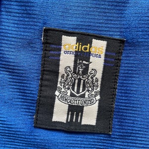 K&ouml;nnte beinhalten: Ein schwarz-wei&szlig;es Patch mit dem Adidas-Logo und dem Text "Official Replica" &uuml;ber dem Wappen von Newcastle United. Das Patch ist auf einen blauen Stoff gen&auml;ht.