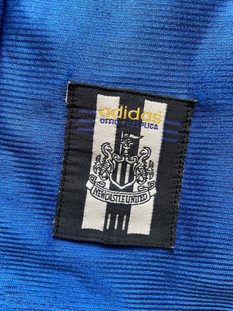K&ouml;nnte beinhalten: Ein schwarz-wei&szlig;es Patch mit dem Adidas-Logo und dem Text "Official Replica" &uuml;ber dem Wappen von Newcastle United. Das Patch ist auf einen blauen Stoff gen&auml;ht.