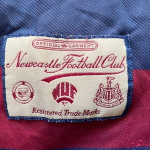 May include: Close-up of a Newcastle Football Club label on a blue and maroon striped fabric. The label features the club's name in cursive script, along with several emblems and the text "Registered Trade Marks."
