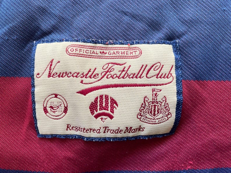 May include: Close-up of a Newcastle Football Club label on a blue and maroon striped fabric. The label features the club's name in cursive script, along with several emblems and the text "Registered Trade Marks."