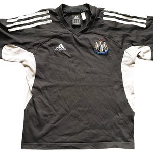 May include: Black and white football shirt with the Adidas logo and the Newcastle United Football Club crest. The shirt has white stripes on the shoulders and sleeves.