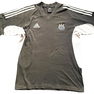 May include: A black and white Adidas t-shirt with a Newcastle United crest on the right side and the Adidas logo on the left side. The shirt has white stripes on the shoulders and sleeves.