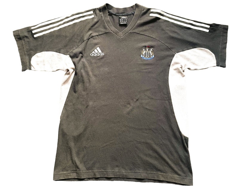 May include: A black and white Adidas t-shirt with a Newcastle United crest on the right side and the Adidas logo on the left side. The shirt has white stripes on the shoulders and sleeves.