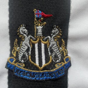 Puede incluir: Parche bordado con el escudo del Newcastle United. Presenta dos caballitos de mar, un escudo con tres franjas verticales y una cinta azul con "NEWCASTLE UNITED". Sobre un fondo a rayas blancas y negras.