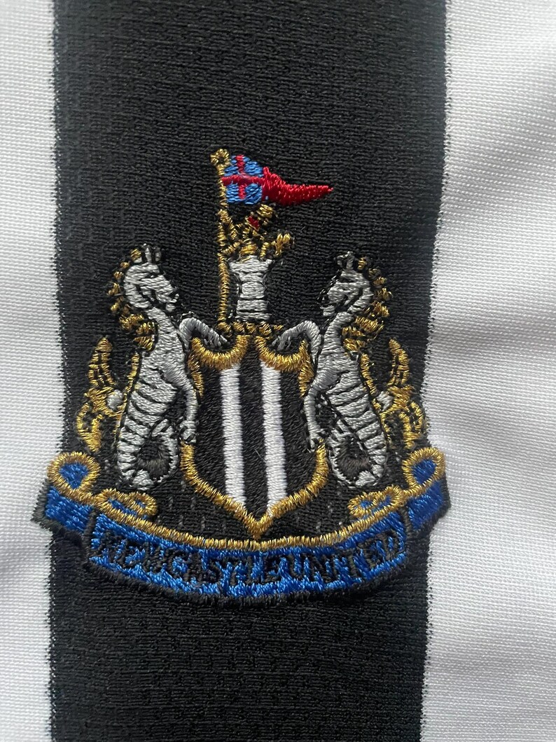 Puede incluir: Parche bordado con el escudo del Newcastle United. Presenta dos caballitos de mar, un escudo con tres franjas verticales y una cinta azul con "NEWCASTLE UNITED". Sobre un fondo a rayas blancas y negras.