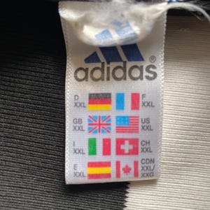 Puede incluir: Etiqueta blanca con texto negro que dice "adidas" y un logotipo de adidas en blanco y negro. La etiqueta tambi&eacute;n tiene una tabla de tallas con diferentes abreviaturas de tallas de pa&iacute;ses y banderas. La etiqueta est&aacute; sobre un fondo a rayas blanco y negro.