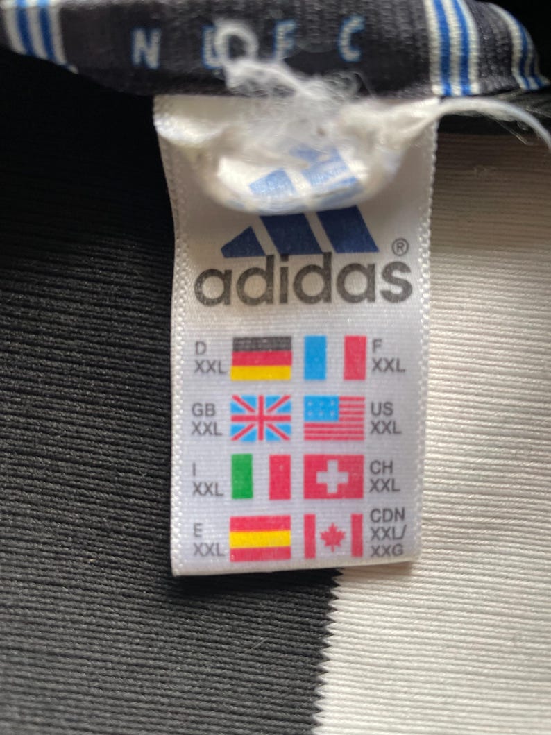 Puede incluir: Etiqueta blanca con texto negro que dice "adidas" y un logotipo de adidas en blanco y negro. La etiqueta tambi&eacute;n tiene una tabla de tallas con diferentes abreviaturas de tallas de pa&iacute;ses y banderas. La etiqueta est&aacute; sobre un fondo a rayas blanco y negro.