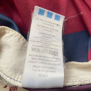 May include: A close-up of a garment label, predominantly white with blue text and a blue logo. The label reads "DIE WELTMARKE MIT DEN 3 STREIFEN" and "THE BRAND WITH THE 3 STRIPES". It also states "Made in England".