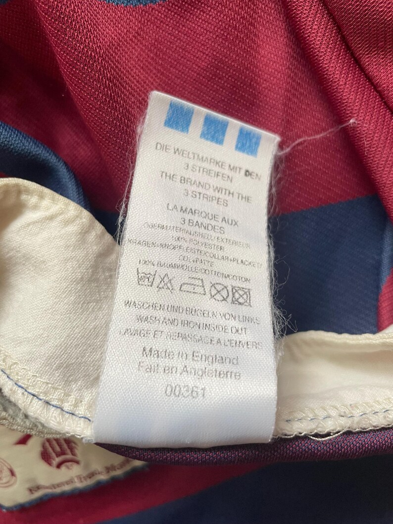 May include: A close-up of a garment label, predominantly white with blue text and a blue logo. The label reads "DIE WELTMARKE MIT DEN 3 STREIFEN" and "THE BRAND WITH THE 3 STRIPES". It also states "Made in England".