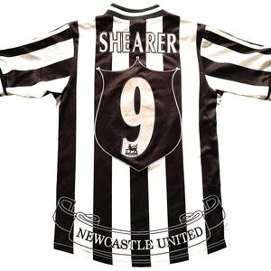 May include: A black and white striped Newcastle United soccer jersey. The jersey features the name "SHEARER" and the number "9" on the back. The team name "NEWCASTLE UNITED" is displayed on a banner at the bottom.