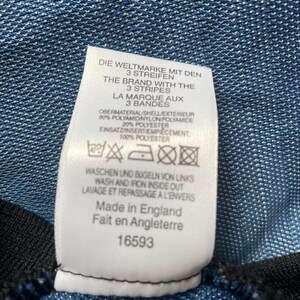 May include: Close-up of a white clothing tag with text in multiple languages, including English, French, and German. The tag includes washing instructions and the phrase "Made in England". The background is a textured blue fabric.