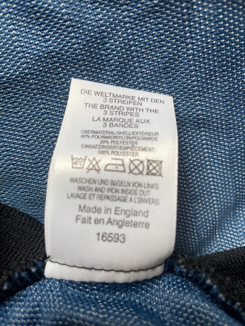 May include: Close-up of a white clothing tag with text in multiple languages, including English, French, and German. The tag includes washing instructions and the phrase "Made in England". The background is a textured blue fabric.