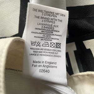 May include: Close-up of a white tag with text in multiple languages, including English, German, and French. The tag lists fabric composition as 70% cotton and 30% polyester. The text also includes washing instructions and the phrase "Made in England."