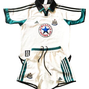 Pu&ograve; includere: Completo da calcio bianco con dettagli neri e verdi. La maglia ha il logo Adidas, il logo Newcastle Brown Ale e lo stemma della squadra. I pantaloncini hanno il logo Adidas e lo stemma della squadra.