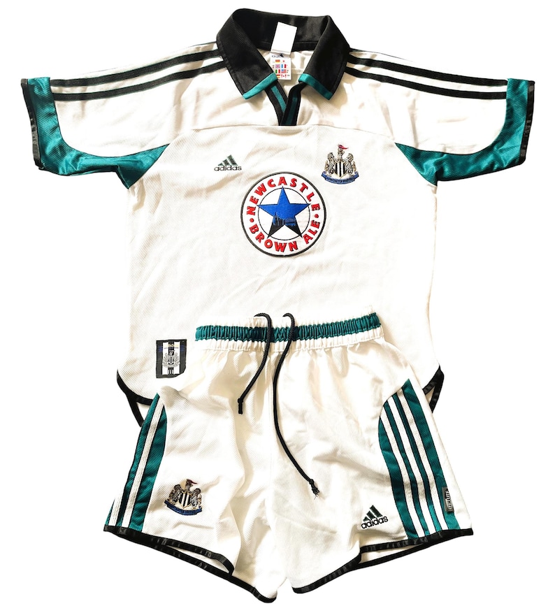 Pu&ograve; includere: Completo da calcio bianco con dettagli neri e verdi. La maglia ha il logo Adidas, il logo Newcastle Brown Ale e lo stemma della squadra. I pantaloncini hanno il logo Adidas e lo stemma della squadra.