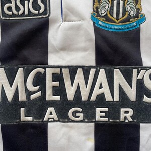 May include: Close-up of a vintage Newcastle United football jersey. The jersey features black and white vertical stripes, with the McEwan's Lager logo prominently displayed. The Asics logo and team crest are also visible.