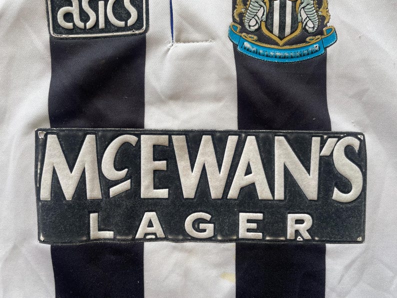 May include: Close-up of a vintage Newcastle United football jersey. The jersey features black and white vertical stripes, with the McEwan's Lager logo prominently displayed. The Asics logo and team crest are also visible.