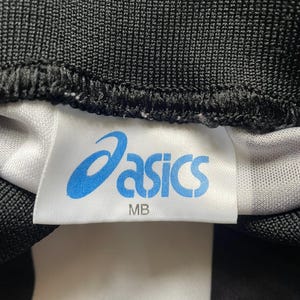 K&ouml;nnte beinhalten: Nahaufnahme eines wei&szlig;en Etiketts mit dem blauen "asics"-Logo und dem Text "MB". Das Etikett ist auf einen schwarz-wei&szlig; gestreiften Stoff gen&auml;ht. Der Stoff hat ein strukturiertes Aussehen.