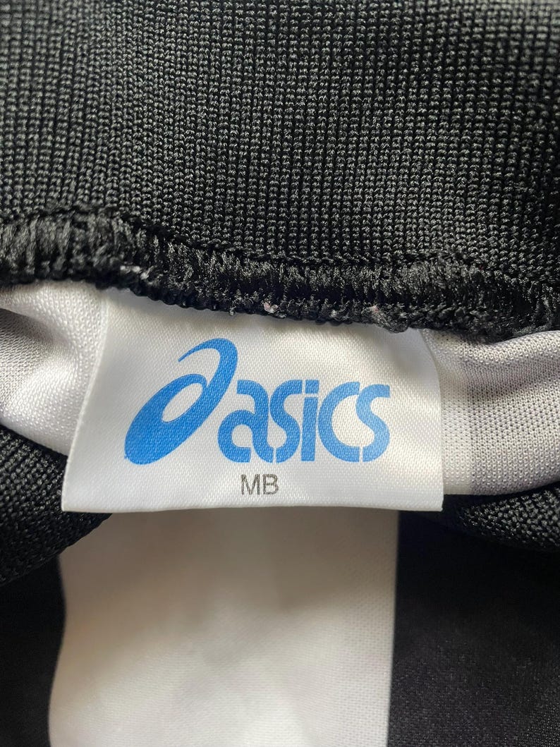 K&ouml;nnte beinhalten: Nahaufnahme eines wei&szlig;en Etiketts mit dem blauen "asics"-Logo und dem Text "MB". Das Etikett ist auf einen schwarz-wei&szlig; gestreiften Stoff gen&auml;ht. Der Stoff hat ein strukturiertes Aussehen.