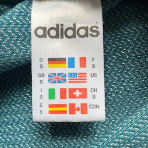 Pu&ograve; includere: Un'etichetta Adidas bianca con logo e bandiere di paesi, tra cui Germania, Francia e Regno Unito. Lo sfondo &egrave; un tessuto turchese con trama.