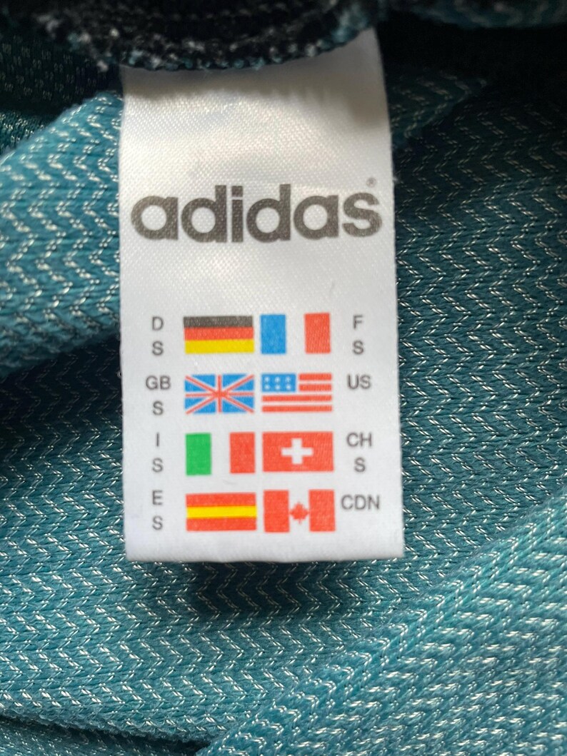 Pu&ograve; includere: Un'etichetta Adidas bianca con logo e bandiere di paesi, tra cui Germania, Francia e Regno Unito. Lo sfondo &egrave; un tessuto turchese con trama.