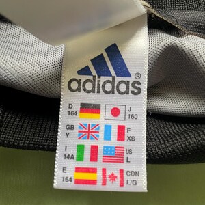 Pu&ograve; includere: Primo piano di un'etichetta Adidas con il logo del marchio e varie bandiere nazionali. L'etichetta include informazioni sulle taglie e conversioni di taglie internazionali. Lo sfondo &egrave; un tessuto grigio strutturato.