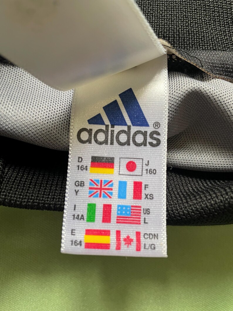 Pu&ograve; includere: Primo piano di un'etichetta Adidas con il logo del marchio e varie bandiere nazionali. L'etichetta include informazioni sulle taglie e conversioni di taglie internazionali. Lo sfondo &egrave; un tessuto grigio strutturato.