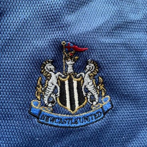 Op de afbeelding: Close-up van een blauwe stof met het geborduurde embleem van Newcastle United. Het embleem toont een schild met een zwart-wit ontwerp, geflankeerd door twee zeepaardjes, en de woorden "Newcastle United" in blauw.