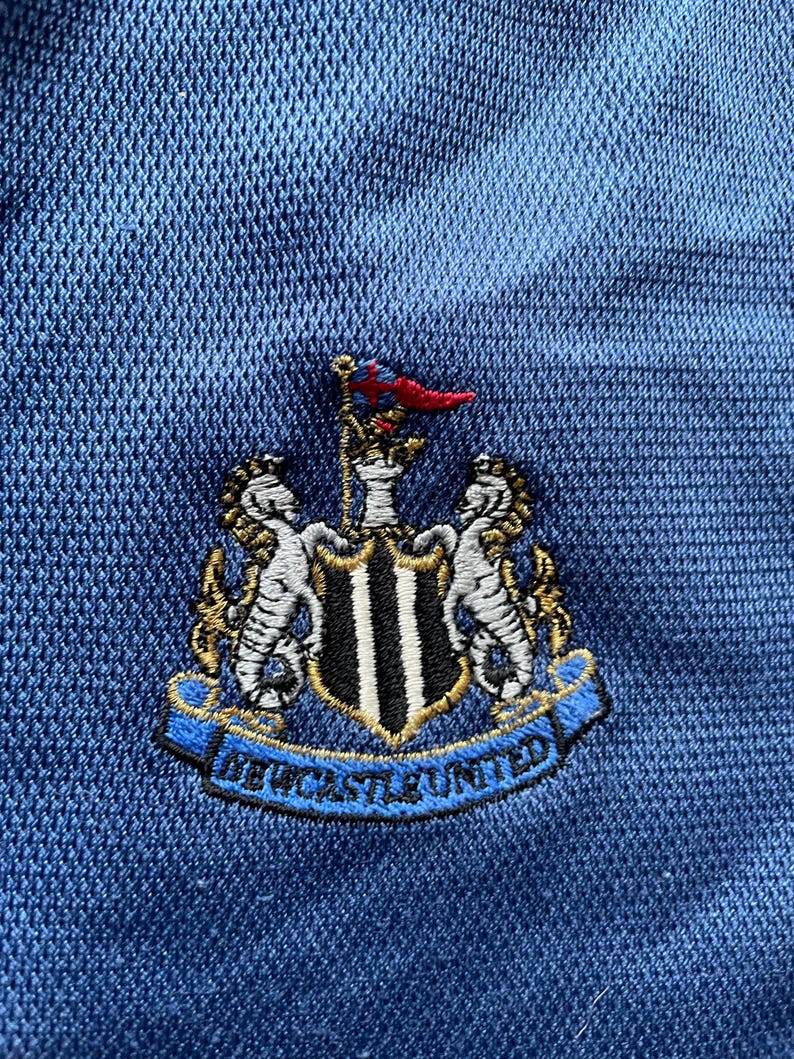 Op de afbeelding: Close-up van een blauwe stof met het geborduurde embleem van Newcastle United. Het embleem toont een schild met een zwart-wit ontwerp, geflankeerd door twee zeepaardjes, en de woorden "Newcastle United" in blauw.