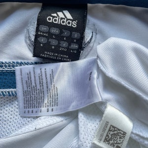 May include: White Adidas shirt with a blue trim and a size tag that reads "UK USA 44/46 L 6 8 J CDM 6 8 2XO LIG MADE IN CHINA FABRIQUE EN CHINE".