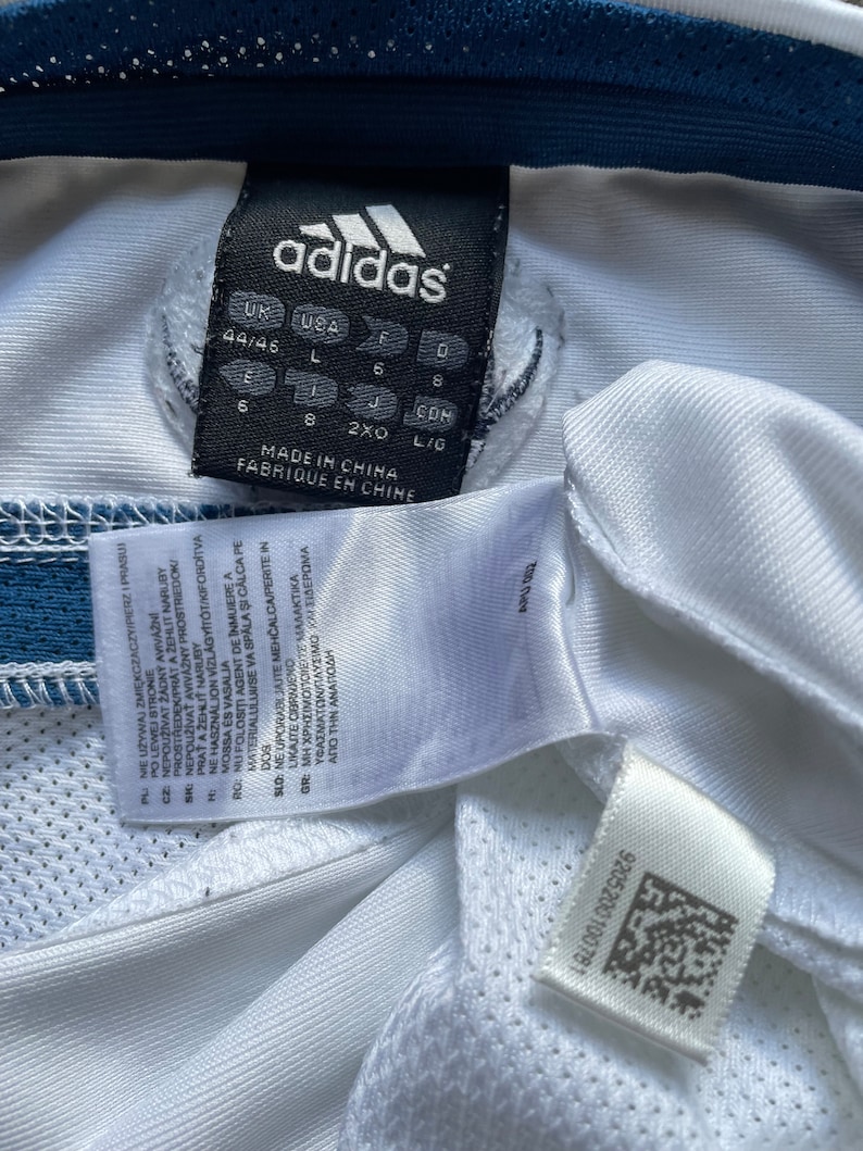 May include: White Adidas shirt with a blue trim and a size tag that reads "UK USA 44/46 L 6 8 J CDM 6 8 2XO LIG MADE IN CHINA FABRIQUE EN CHINE".
