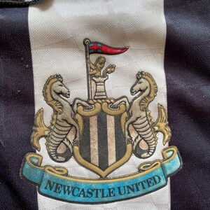 Pode incluir: Uma camisa de futebol preta e branca com o bras&atilde;o do Newcastle United na frente. O bras&atilde;o apresenta um escudo com listras pretas e brancas, dois cavalos-marinhos e uma faixa que diz "Newcastle United".
