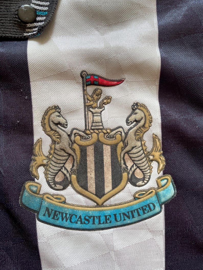 Pode incluir: Uma camisa de futebol preta e branca com o bras&atilde;o do Newcastle United na frente. O bras&atilde;o apresenta um escudo com listras pretas e brancas, dois cavalos-marinhos e uma faixa que diz "Newcastle United".