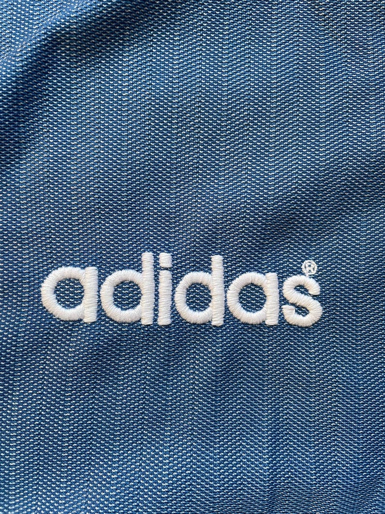 May include: A close-up of a blue fabric with the word "adidas" embroidered in white.