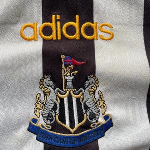 May include: Close-up of a black and white striped jersey with the Adidas logo in gold. The Newcastle United crest is embroidered below, featuring two seahorses and a shield with the text "NEWCASTLE UNITED" on a blue banner.
