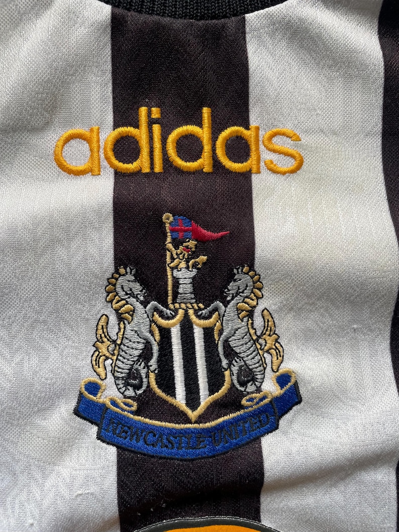 May include: Close-up of a black and white striped jersey with the Adidas logo in gold. The Newcastle United crest is embroidered below, featuring two seahorses and a shield with the text "NEWCASTLE UNITED" on a blue banner.