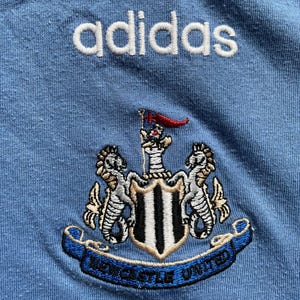 May include: A light blue t-shirt with the adidas logo embroidered in white and the Newcastle United crest embroidered in black, white, and gold.