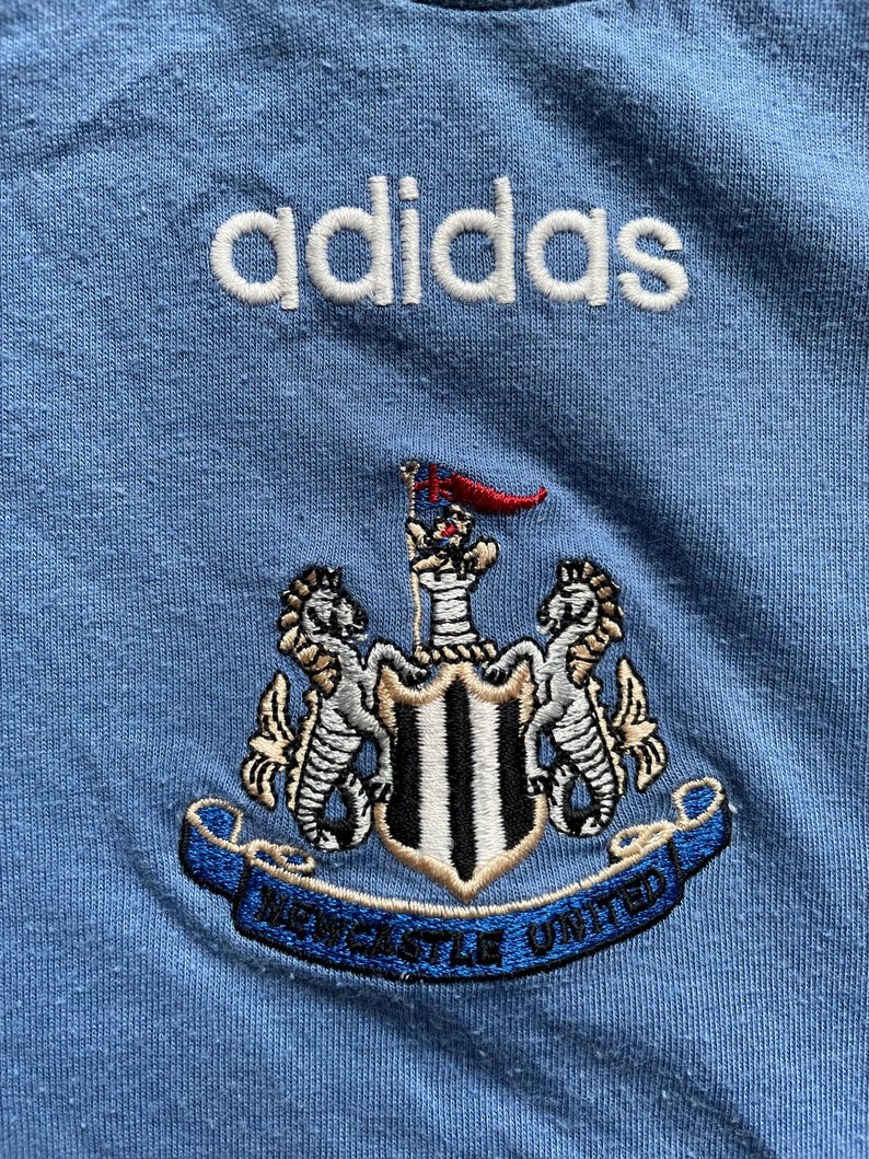May include: A light blue t-shirt with the adidas logo embroidered in white and the Newcastle United crest embroidered in black, white, and gold.