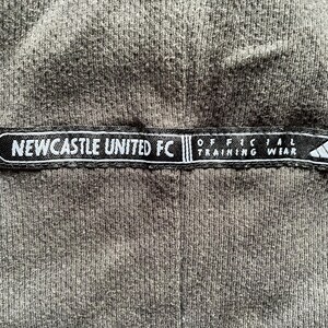 May include: Black and white label with the text "NEWCASTLE UNITED FC" and "OFFICIAL TRAINING WEAR" on a gray fabric background.