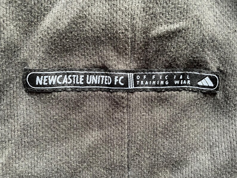 May include: Black and white label with the text "NEWCASTLE UNITED FC" and "OFFICIAL TRAINING WEAR" on a gray fabric background.