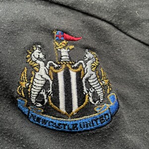 May include: Black embroidered patch with a crest featuring a shield with black and white stripes, two seahorses, and a flag. The text "NEWCASTLE UNITED" is written on a blue ribbon.