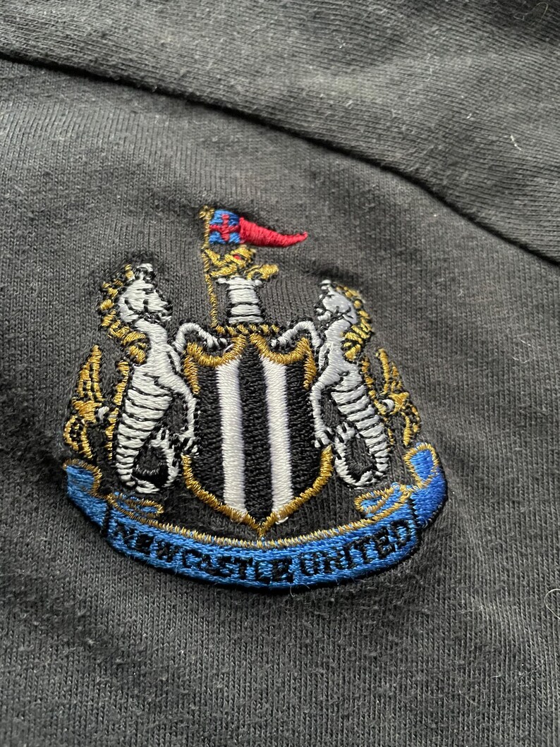May include: Black embroidered patch with a crest featuring a shield with black and white stripes, two seahorses, and a flag. The text "NEWCASTLE UNITED" is written on a blue ribbon.