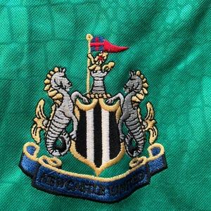 May include: Close-up of a green fabric with the embroidered Newcastle United crest. The crest features two seahorses, a shield with black and white stripes, and a banner with the text "NEWCASTLE UNITED".
