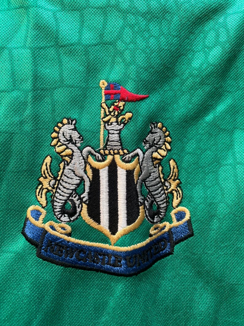 May include: Close-up of a green fabric with the embroidered Newcastle United crest. The crest features two seahorses, a shield with black and white stripes, and a banner with the text "NEWCASTLE UNITED".