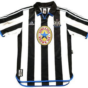 May include: Black and white striped soccer jersey with blue accents. Features the Adidas logo, a Newcastle United crest, and a circular emblem with text and a blue star. The jersey has short sleeves.